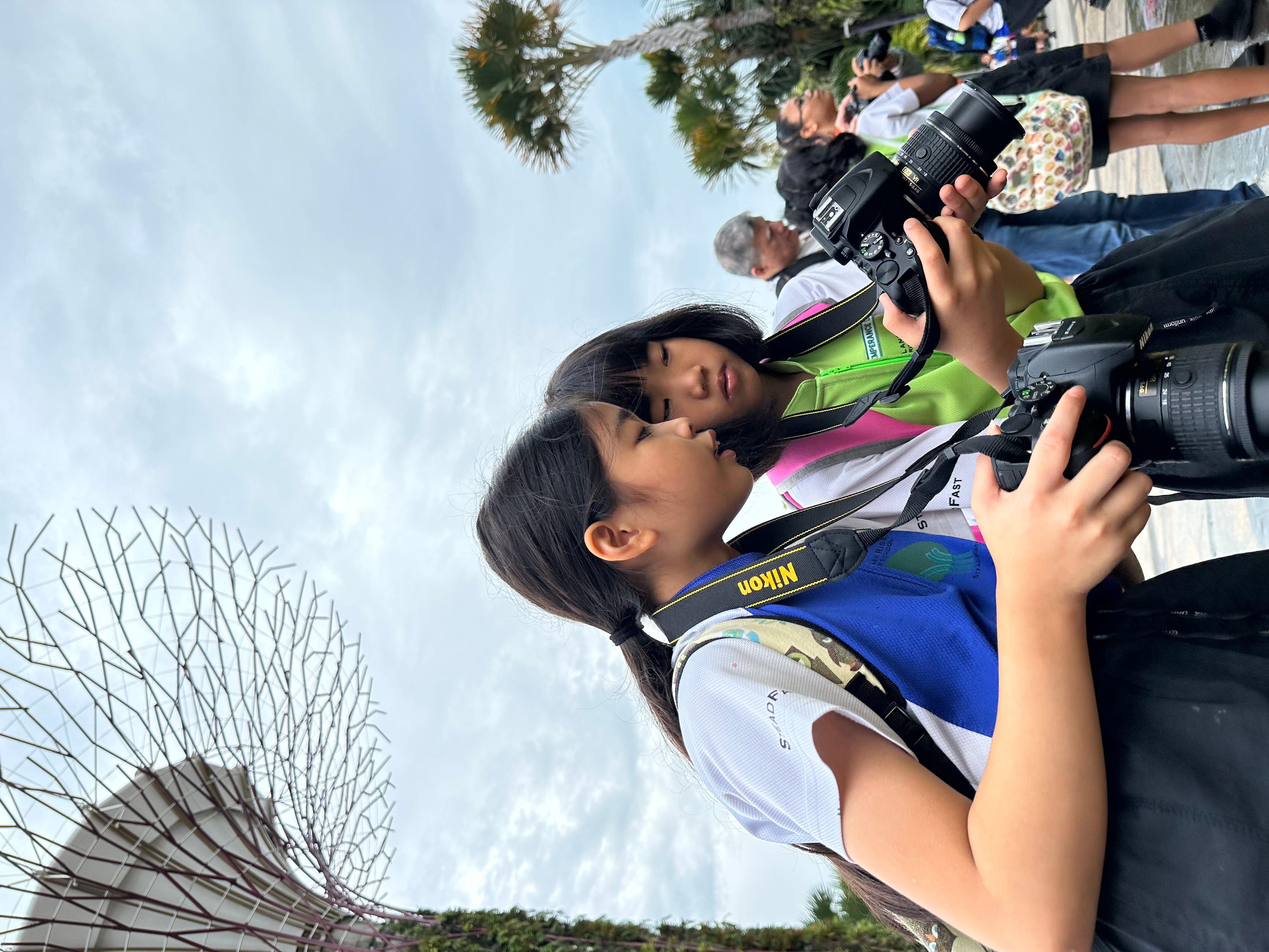 Two young girls review photos on Nikon DSLR cameras outdoors near tall tree-like structures under a cloudy sky.